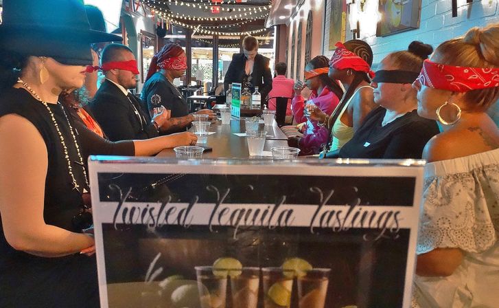 Blindfolded adults lined up along a long bar under string lights, sampling shots and guided by a server during a lively indoor tequila tasting event.