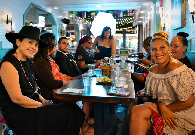 Smiling group dining at a lively neighborhood restaurant bar — long wooden table with a tequila bottle, glasses and a plate of limes, string lights and international flags overhead, casual evening gathering.