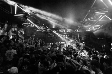 Black-and-white photo of a packed indoor nightclub dance floor with geometric LED ceiling lights, smoke beams and a lively crowd dancing.