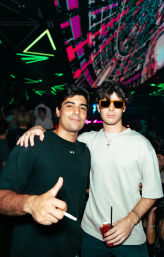 Two young men posing in a neon-lit nightclub under colorful LED ceiling visuals — one wearing orange sunglasses holding a red cocktail, the other giving a thumbs-up and holding a cigarette, urban nightlife scene.