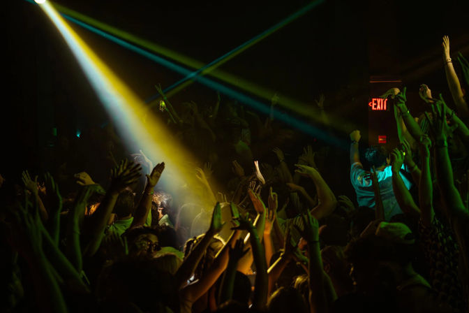 Energetic concert crowd in a dark music venue, hands raised under green and yellow spotlights with crisscrossing beams.