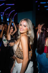 Smiling young woman in a beige crop top and white pants on a crowded nightclub dance floor, blonde hair and tan patterned shoulder bag in a vibrant nightlife scene.