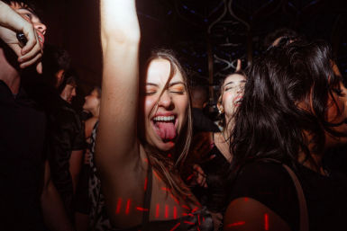 Energetic young woman on a crowded nightclub dance floor, arm raised and tongue out, eyes closed amid friends and red laser lights.