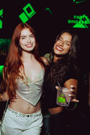Two smiling women posing in a neon-lit nightclub — red-haired woman in a metallic crop top and white jeans beside a dark-haired woman in black holding a cocktail, lively city nightlife vibe.
