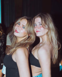Close-up portrait of two young women in black tops posing back-to-back and smiling at a city nightlife party, warm flash lighting and candid party vibe