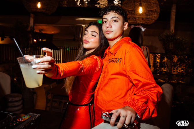 Two partygoers in bright orange outfits at a lively nightclub lounge, woman holding a cocktail toward the camera while a man poses near a DJ booth under warm hanging lights.