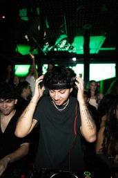 Tattooed DJ adjusting headphones at a packed nightclub DJ booth with green LED screens and dancing crowd