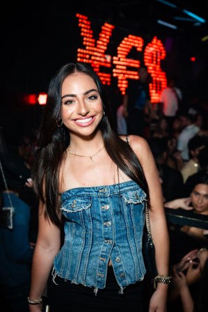Smiling woman in a denim corset top at a lively nightclub, red LED currency symbols glowing behind her amid a crowded party scene