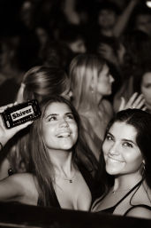 Black-and-white nightclub photo of two smiling young women on the dance floor, one holding a phone displaying 'Shiver,' blurred party crowd in background.