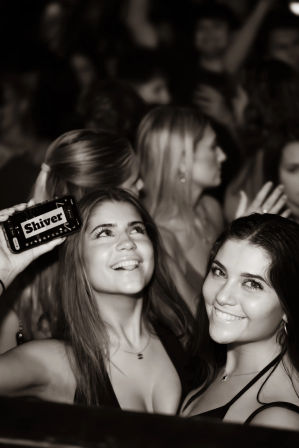 Black-and-white nightclub photo of two smiling young women on the dance floor, one holding a phone displaying 'Shiver,' blurred party crowd in background.