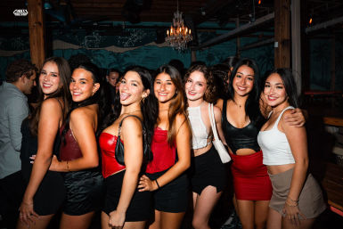 Group of seven friends posing and smiling in a lively nightclub party scene — chic outfits, colorful lighting, chandelier and wood-beamed interior, perfect for a girls' night out.