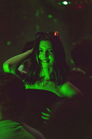 Smiling woman enjoying a crowded nightclub nightlife scene, bathed in neon green light with sunglasses on her head and a pendant necklace visible