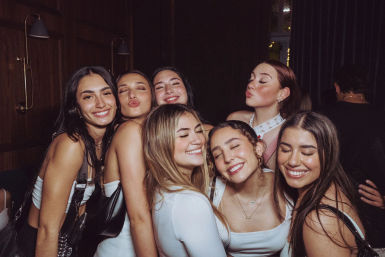 Seven young women smiling and posing closely in an indoor lounge during a night out, dressed up and enjoying a group celebration