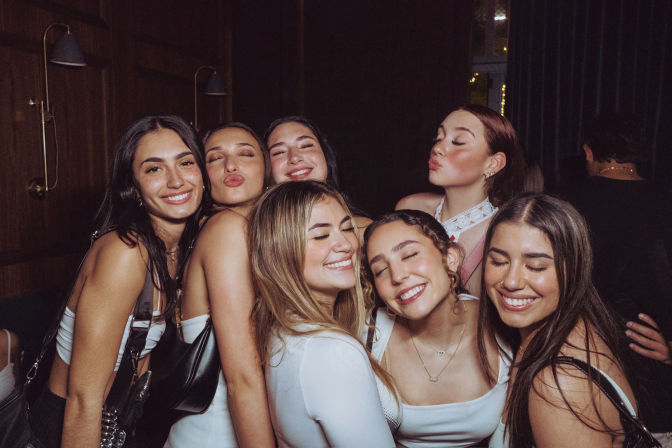 Seven young women smiling and posing closely in an indoor lounge during a night out, dressed up and enjoying a group celebration