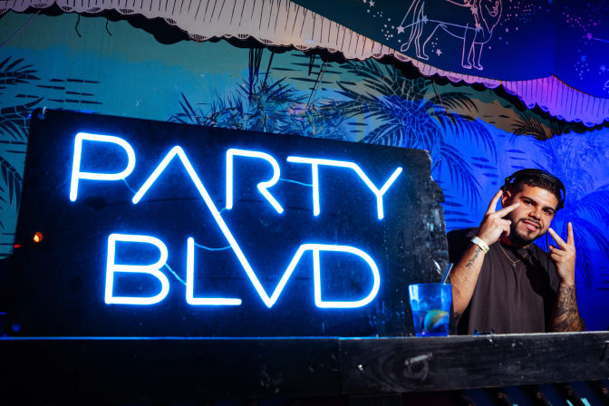 Blue neon "PARTY BLVD" sign over a nightclub DJ booth, smiling DJ in headphones flashing peace signs beside a cocktail glass against a tropical mural backdrop.