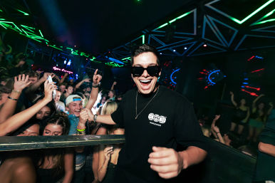 Smiling man in sunglasses and black shirt posing at a crowded nightclub dance floor with cheering young adults, neon geometric lights, and energetic party atmosphere.
