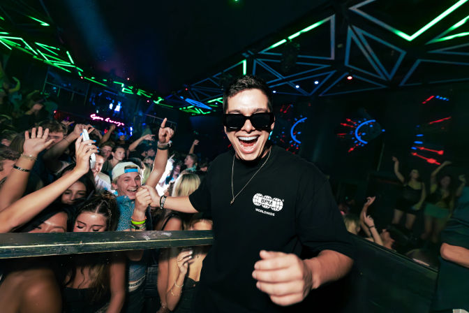 Smiling man in sunglasses and black shirt posing at a crowded nightclub dance floor with cheering young adults, neon geometric lights, and energetic party atmosphere.