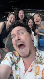 Energetic nighttime outdoor selfie of five laughing people — a man in a tropical-print shirt in the foreground and four smiling females behind him, candid group photo vibe.