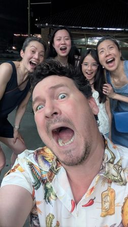 Energetic nighttime outdoor selfie of five laughing people — a man in a tropical-print shirt in the foreground and four smiling females behind him, candid group photo vibe.