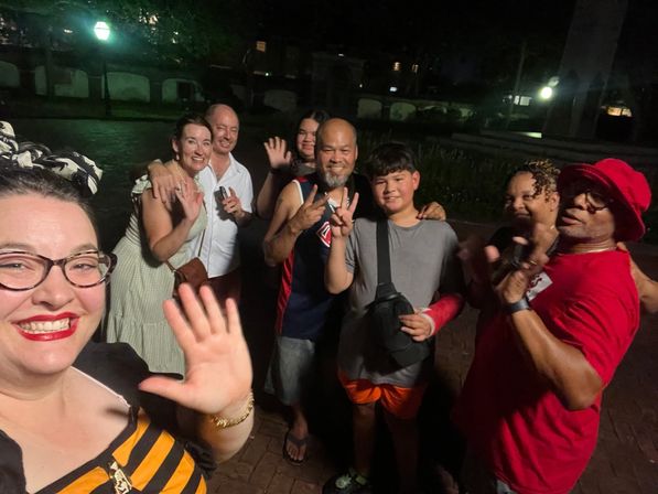 Happy group selfie of eight people smiling, waving and flashing peace signs on a dimly lit urban park walkway at night with lamp posts in the background