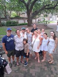 Smiling multigenerational family posing on a brick walkway under a large oak tree in a shady historic courtyard with benches and a lamp post