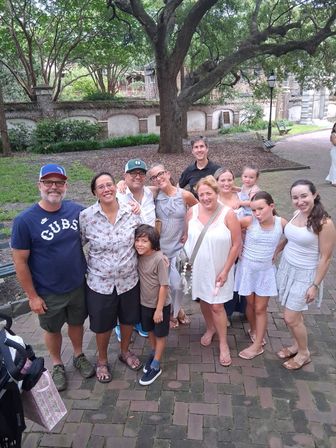 Smiling multigenerational family posing on a brick walkway under a large oak tree in a shady historic courtyard with benches and a lamp post