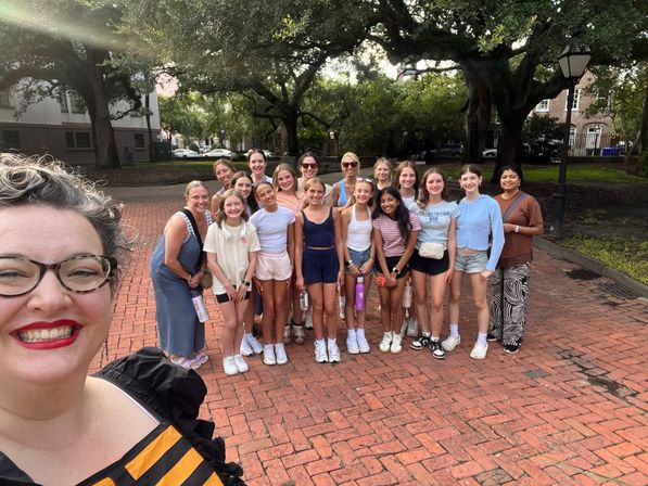 Selfie of a smiling woman with a group of teen girls and adult chaperones standing on a sunlit historic brick plaza under large oak trees in a city park.