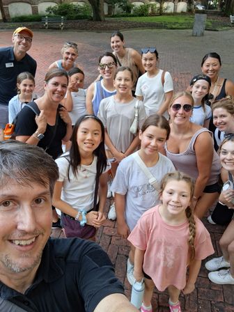 Group selfie of smiling adults and children in casual summer clothes in a shaded brick courtyard with trees and benches