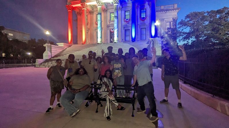 Group of people posing and waving at night on wide stone steps in front of a neoclassical civic building with red, white and blue illuminated columns — playful city outing.