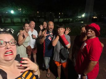 Nighttime outdoor plaza group selfie of eight people making surprised, playful faces — woman with red lipstick in foreground, boy with red arm cast covering his mouth, man in red hat, lampposts and trees in the background.