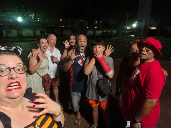 Nighttime outdoor plaza group selfie of eight people making surprised, playful faces — woman with red lipstick in foreground, boy with red arm cast covering his mouth, man in red hat, lampposts and trees in the background.