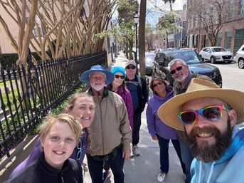 Smiling group selfie of nine on a sunny spring walking tour down a historic city sidewalk lined with iron fences, crepe myrtle trees, brick rowhouses and parked cars.