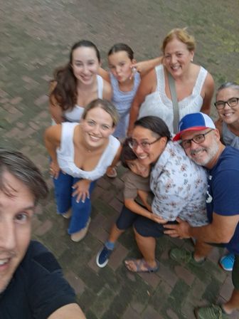 Overhead selfie of a smiling multi-generation group of adults and children huddled on a brick sidewalk outdoors, wearing casual summer clothes