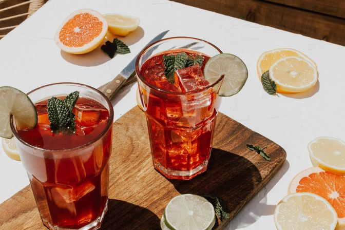 Sunlit outdoor scene with two glasses of iced citrus tea on a wooden board, garnished with lime slices, mint and ice cubes, surrounded by grapefruit and lemon slices on a white table — refreshing summer drink.