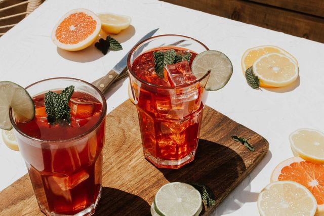 Sunlit outdoor scene with two glasses of iced citrus tea on a wooden board, garnished with lime slices, mint and ice cubes, surrounded by grapefruit and lemon slices on a white table — refreshing summer drink.