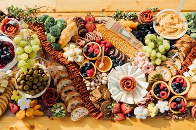 Vibrant charcuterie grazing board on a wooden table with assorted cheeses, crackers, sliced baguette and croissants, salami roses, green grapes, mixed berries, olives, nuts, dried apricots, honey and creamy dip.
