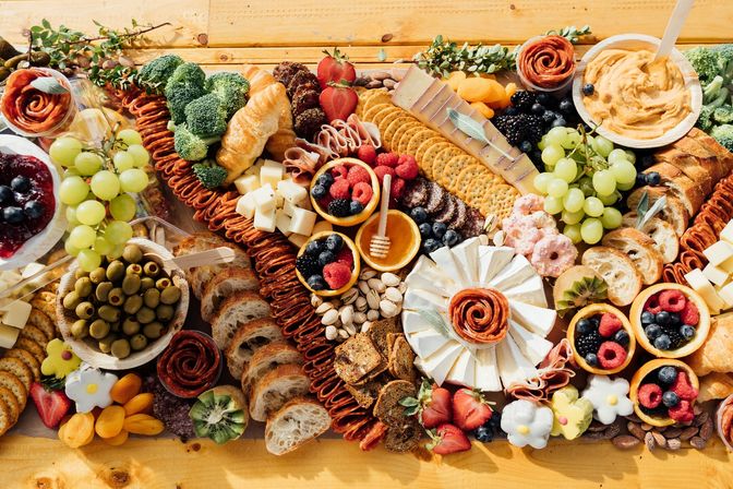 Vibrant charcuterie grazing board on a wooden table with assorted cheeses, crackers, sliced baguette and croissants, salami roses, green grapes, mixed berries, olives, nuts, dried apricots, honey and creamy dip.