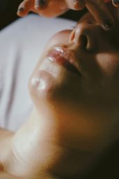 Close-up portrait of a lower face showing glossy lips, chin and nostrils with fingers hovering above, warm soft lighting highlighting skin texture.