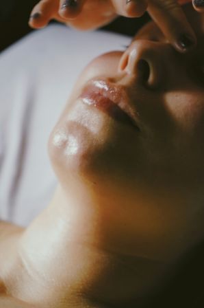 Close-up portrait of a lower face showing glossy lips, chin and nostrils with fingers hovering above, warm soft lighting highlighting skin texture.