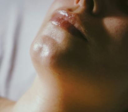 Close-up portrait of a lower face showing glossy lips, chin and nostrils with fingers hovering above, warm soft lighting highlighting skin texture.