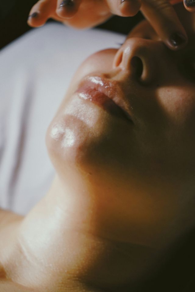 Close-up portrait of a lower face showing glossy lips, chin and nostrils with fingers hovering above, warm soft lighting highlighting skin texture.