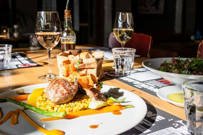 Sunlit restaurant table with a gourmet plated lamb chop on pearl couscous, asparagus and carrot ribbons with a crispy garnish, accompanied by wine glasses, a bread basket and salad — fine dining scene.