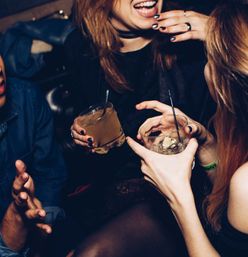 Close-up of friends holding cocktails and ice, laughing at a lively bar — nightlife scene with rings, dark nail polish and wristband.