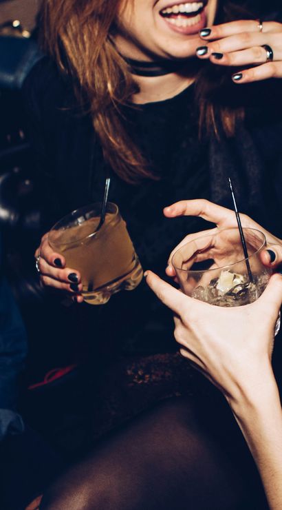 Close-up of friends holding cocktails and ice, laughing at a lively bar — nightlife scene with rings, dark nail polish and wristband.