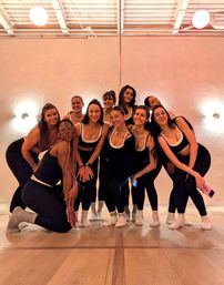 Smiling women in matching black-and-white workout outfits posing together on a wooden floor around a center pole in a pink-walled dance/pole fitness studio.