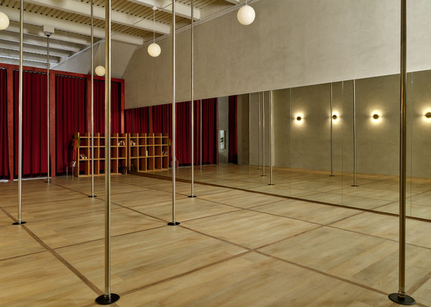 Bright pole fitness studio with rows of brass poles, full-length mirrored wall, light wood floor, red stage curtains, globe pendant lights and cubby storage.