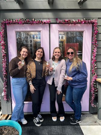 Four smiling women holding small perfume bottles in front of a pink double-door boutique entrance decorated with pink floral garland, casual outdoor group photo.
