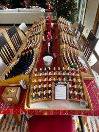 Festive dining table set up for a scent-blending workshop: dozens of colorful dropper bottles neatly arranged in wooden display racks along a red holiday runner, with clipboards, cups, candles and a Christmas tree in the living room background.
