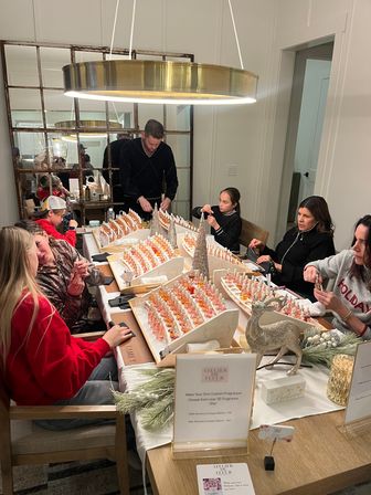 Family and friends gathered around a modern dining table making custom fragrances at a holiday workshop, rows of small glass scent bottles, festive greenery and a gold pendant light