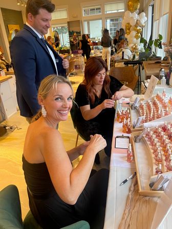 Smiling woman at a boutique salon event sampling dropper skincare serums from a tabletop display while an attendant fills bottles, with gold-and-white balloons and salon interior in the background.